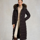 Woman wearing a long dark brown puffer coat with a fur-lined hood on a plain background